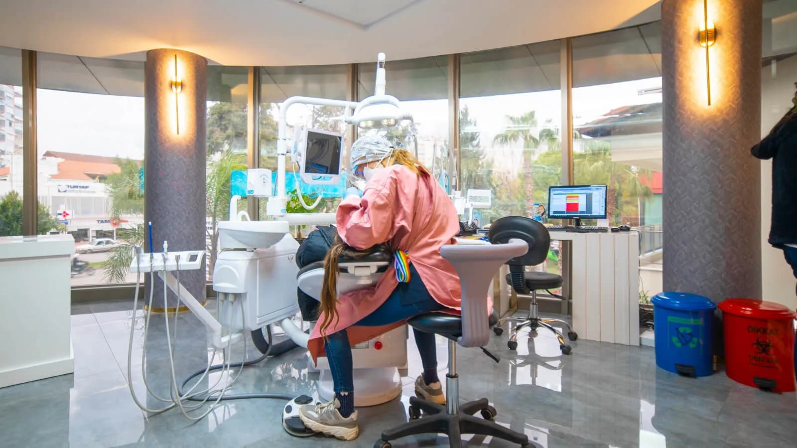 A masked clinician at Ballıpınar Dental treating a patient in a window-lit surgery, both seen from behind
